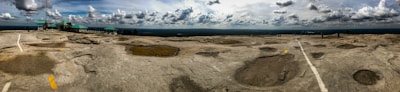 A vast rocky terrain stretches across the image with distinctive craters and a few painted markings. The sky above is partly cloudy, casting dynamic shadows on the ground. Structures with teal roofs are visible on the left side, likely facilities or observation points.