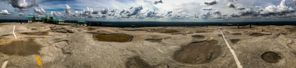 A vast rocky terrain stretches across the image with distinctive craters and a few painted markings. The sky above is partly cloudy, casting dynamic shadows on the ground. Structures with teal roofs are visible on the left side, likely facilities or observation points.