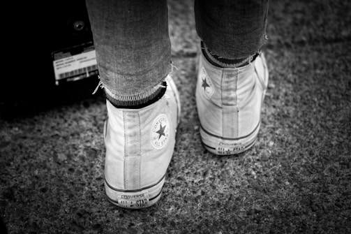 A black and white image showing the back of a pair of worn high-top Converse All Star sneakers. The shoes are on a textured surfaced, possibly pavement, and the wearer is dressed in fitted pants.