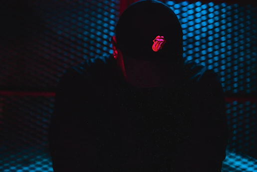 A person wearing a cap with a prominent red tongue and lips logo is silhouetted against a textured, metallic background. The scene is dimly lit with a blue hue, creating a moody and mysterious atmosphere.