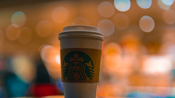 A white cup with a coffee brand logo is in focus against a blurred background of warm, yellow and orange bokeh lights.
