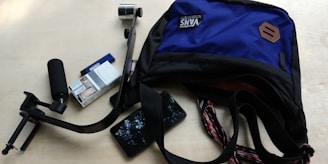 A blue and black backpack partially open with a stabilizer, a pack of cigarettes, a lighter, and a smartphone scattered around it on a light wooden surface.