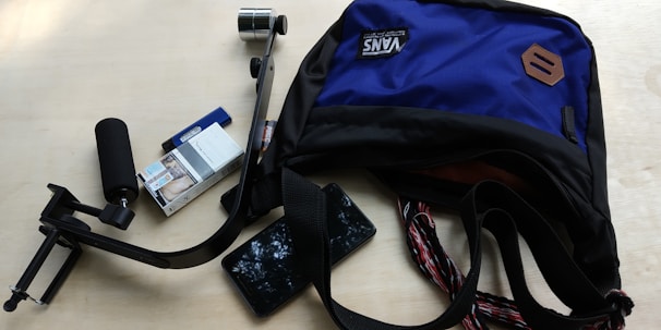 A blue and black backpack partially open with a stabilizer, a pack of cigarettes, a lighter, and a smartphone scattered around it on a light wooden surface.