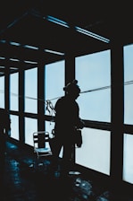 A silhouette of a person wearing a harness is seen standing near a large floor-to-ceiling window. The figure appears to be engaged in an activity related to window cleaning or maintenance, with equipment attached to their body. The overall setting is dimly lit with shadows cast on the floor.