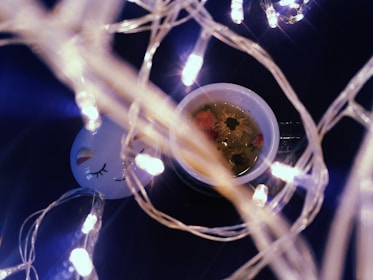 Delicate string lights wrapped around a white ceramic mug filled with floral tea, featuring visible flower buds. A serene face design with closed eyes is on a nearby object, contributing to a cozy atmosphere.