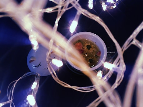 Delicate string lights wrapped around a white ceramic mug filled with floral tea, featuring visible flower buds. A serene face design with closed eyes is on a nearby object, contributing to a cozy atmosphere.