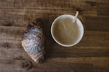 A steaming cup of coffee beside a croissant on a rustic table.