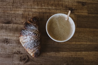 A cup of coffee with frothy milk is placed beside a croissant dusted with powdered sugar on a wooden table. The croissant appears slightly burnt on the edges, and a small wooden stirrer is placed in the cup.