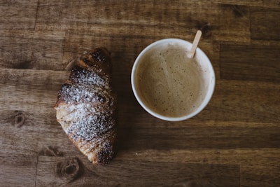 A cup of coffee with frothy milk is placed beside a croissant dusted with powdered sugar on a wooden table. The croissant appears slightly burnt on the edges, and a small wooden stirrer is placed in the cup.