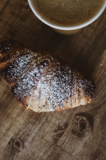 A freshly baked golden croissant on a rustic wooden table with a cup of coffee beside it.