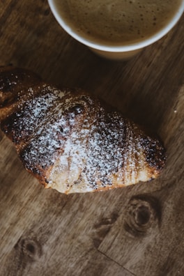Close-up of a flaky croissant resting beside a steaming cup of specialty coffee on a rustic wooden table.