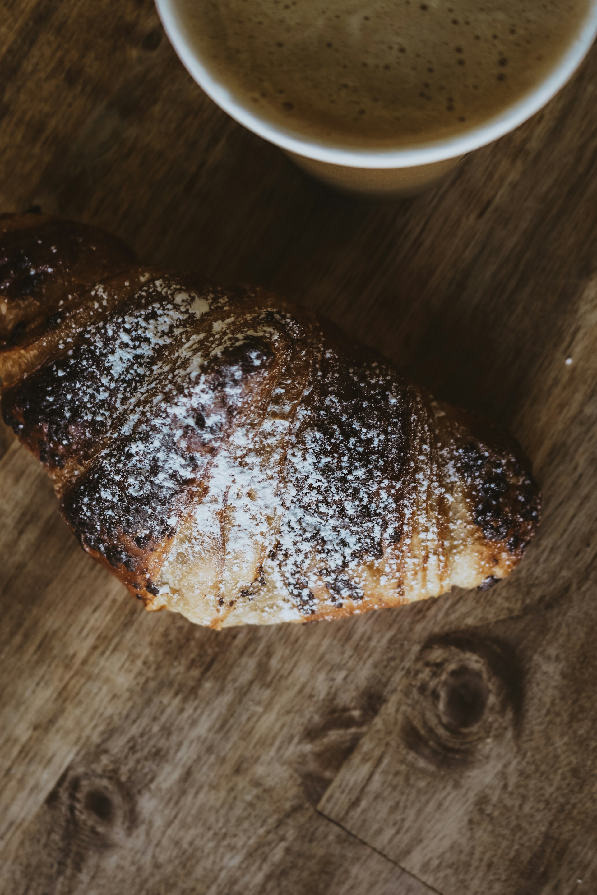 A freshly baked croissant, golden and flaky, placed on a rustic wooden table with a cup of steaming coffee beside it.