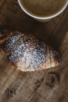 A perfectly baked croissant rests on a textured wooden surface, lightly dusted with powdered sugar. Beside it, a cup of frothy coffee sits on the table, creating a cozy and inviting breakfast scene.