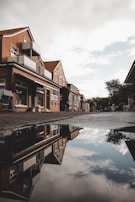 Residential street with brick houses reflecting in a water puddle. The buildings have modern balconies and large windows, with some featuring small storefronts. The sky is partly cloudy, and trees are visible in the background.