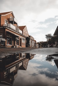 Residential street with brick houses reflecting in a water puddle. The buildings have modern balconies and large windows, with some featuring small storefronts. The sky is partly cloudy, and trees are visible in the background.