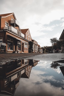 Residential street with brick houses reflecting in a water puddle. The buildings have modern balconies and large windows, with some featuring small storefronts. The sky is partly cloudy, and trees are visible in the background.