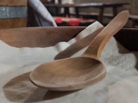A wooden spoon rests on a soft, light-colored cloth. The spoon is made of smooth wood and has a simple, rustic appearance. In the background, there are hints of a wooden table and a barrel, giving the setting a cozy, rustic atmosphere.