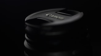 A close-up of a Canon camera lens cap set against a dark background, highlighting the brand logo and circular shape.