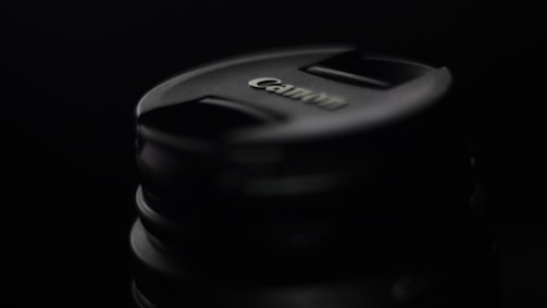A close-up of a Canon camera lens cap set against a dark background, highlighting the brand logo and circular shape.