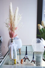 A glass vase with delicate dried ornamental grass and a white ribbon stands next to small figurines of a giraffe, cow, and fox. Several miniature Eiffel Tower replicas are displayed on a glass table, accompanied by a plant pot and a tulip in the background.