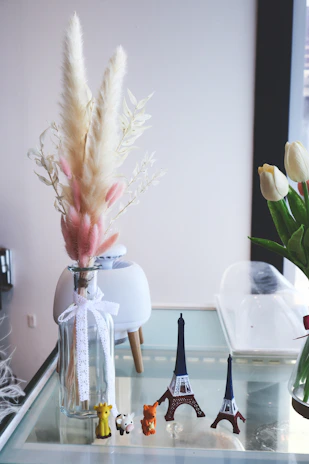 A glass vase with delicate dried ornamental grass and a white ribbon stands next to small figurines of a giraffe, cow, and fox. Several miniature Eiffel Tower replicas are displayed on a glass table, accompanied by a plant pot and a tulip in the background.