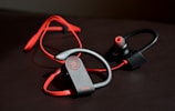A pair of wireless earphones with red and black color accents. The earphones are designed with an ergonomic shape, featuring soft ear hooks for a secure fit. The control panel has a circular red button with a power symbol, and the brand name is in red lettering.