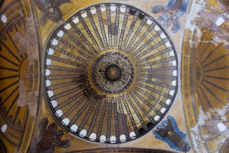 A intricately designed dome featuring concentric patterns with detailed calligraphy at the center. Surrounding the central motif are rows of rectangular shapes with alternating colors, while depictions of ornate figures or angels adorn the surrounding space. The entire structure displays a combination of geometry and artistry.