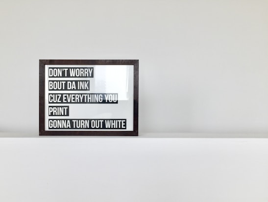 A framed text artwork sits on a white shelf against a light grey wall. The text within the frame is displayed in bold black letters on a white background. The wording is humorous and references printing concerns, stating not to worry about ink.