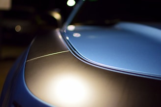 Close-up view of a sleek car hood with a glossy finish, reflecting light. The surface showcases a smooth curvature with a distinct line running through. The background is dark, emphasizing the car's metallic blue shine.