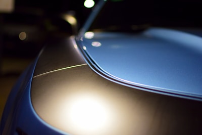 Close-up view of a sleek car hood with a glossy finish, reflecting light. The surface showcases a smooth curvature with a distinct line running through. The background is dark, emphasizing the car's metallic blue shine.