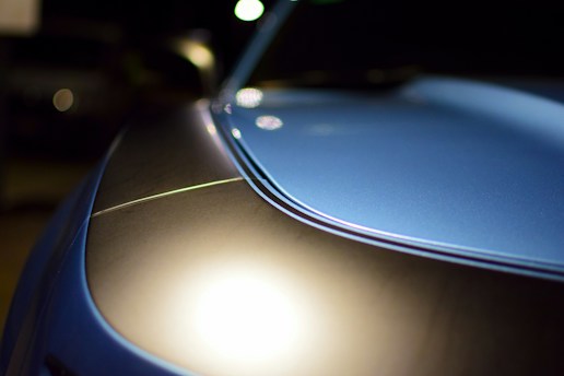 Close-up view of a sleek car hood with a glossy finish, reflecting light. The surface showcases a smooth curvature with a distinct line running through. The background is dark, emphasizing the car's metallic blue shine.