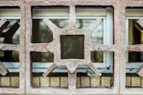 The image captures part of a wall with decorative concrete blocks featuring geometric patterns. Each block has a star-like shape with protruding points surrounding a central square opening. Behind the concrete structure, there are windows, partially covered.