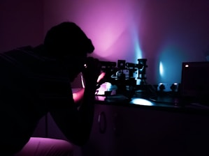 A person is closely examining something using a microscope in a dimly lit room. The ambient lighting creates a striking contrast of purple and blue hues, casting shadows and highlights on the laboratory equipment.
