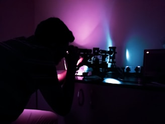 A person is closely examining something using a microscope in a dimly lit room. The ambient lighting creates a striking contrast of purple and blue hues, casting shadows and highlights on the laboratory equipment.