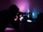 A person is closely examining something using a microscope in a dimly lit room. The ambient lighting creates a striking contrast of purple and blue hues, casting shadows and highlights on the laboratory equipment.