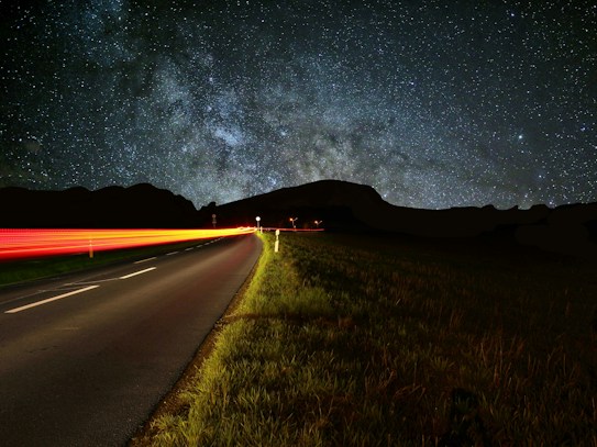 A star-filled night sky stretches across the horizon with a clear view of the Milky Way. A long, empty road runs along the left side, bordered by lush green grass, while a streak of red light trails alongside it, indicating a moving vehicle. The dark silhouette of a hill or mountain is visible against the bright celestial background.