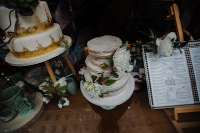 A display of elegantly decorated cakes, with one featuring a golden design and another adorned with white flowers. Nearby, a menu is visible, adding to the aesthetic of a bakery or cafe.