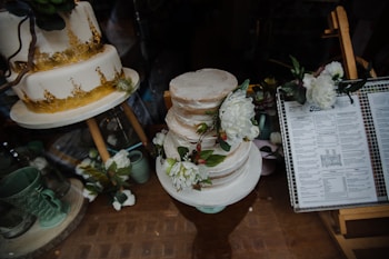 A display of elegantly decorated cakes, with one featuring a golden design and another adorned with white flowers. Nearby, a menu is visible, adding to the aesthetic of a bakery or cafe.