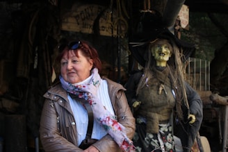 A woman with red hair and glasses on her head is dressed in a brown jacket, white sweater, and a floral scarf. She is smiling and standing in front of a rustic setting that includes a creepy, lifelike witch figure with greenish skin, messy hair, and tattered clothes. The background has various hanging ropes, a wooden sign, and other items suggesting an outdoor or themed environment.