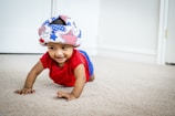 A baby is crawling on a carpeted floor. The baby is wearing a colorful soft helmet with an American flag pattern and a red shirt. They are smiling and appear to be in a bright room with white walls and a door in the background.