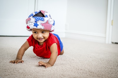 A baby is crawling on a carpeted floor. The baby is wearing a colorful soft helmet with an American flag pattern and a red shirt. They are smiling and appear to be in a bright room with white walls and a door in the background.