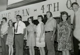 A group of seven people are standing in a line, all dressed in traditional attire from the 1960s or 1970s. The background has text that reads 'GAME PLAN ~ 4TH Q'. The people have a serious demeanor, and they appear to be part of a formal or staged setting. Some individuals are wearing glasses, and the clothing consists of patterned and plain shirts, ties, and dresses.