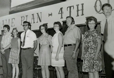 A group of seven people are standing in a line, all dressed in traditional attire from the 1960s or 1970s. The background has text that reads 'GAME PLAN ~ 4TH Q'. The people have a serious demeanor, and they appear to be part of a formal or staged setting. Some individuals are wearing glasses, and the clothing consists of patterned and plain shirts, ties, and dresses.
