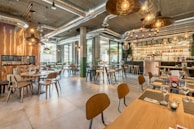A modern restaurant interior with an industrial design style, featuring exposed ductwork and hanging light fixtures. The space includes wooden tables and chairs, a bar area with a variety of bottles displayed, and large windows allowing natural light to enter. The overall look is spacious and inviting, with neatly set tables ready for guests.