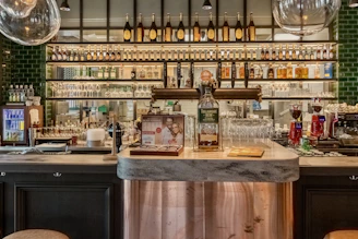 A well-stocked bar features a large assortment of liquor bottles arranged neatly on glass shelves. In front of the shelves, a countertop hosts a prominent bottle of Tullamore Dew whiskey, accompanied by stacked glassware. Multiple taps and coffee machines are present, indicating a diversified beverage offering. The lighting is warm and inviting, highlighting the deep green tiled walls behind.