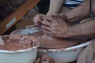 Hands forming clay on a potter's wheel, with wet brown clay being shaped into an object. The scene includes a person assisting or learning pottery, as both sets of hands work together, surrounded by bowls filled with clay. The workspace suggests an artistic and tactile environment.