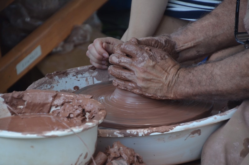 Hands forming clay on a potter's wheel, with wet brown clay being shaped into an object. The scene includes a person assisting or learning pottery, as both sets of hands work together, surrounded by bowls filled with clay. The workspace suggests an artistic and tactile environment.