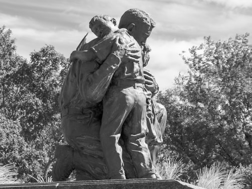 A sculpture depicting an embrace between two figures, likely a parent and child. The figures are captured in a moment of deep connection and emotion, with delicate details in their clothing and posture. Trees and sky form the natural background, adding to the serene atmosphere.