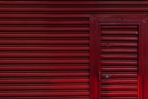 A red corrugated metal garage door with a smaller, matching door integrated into it. The surface is uniform with horizontal ridges and a few visible bolts and handles.