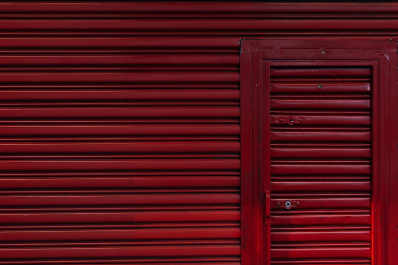 A red corrugated metal garage door with a smaller, matching door integrated into it. The surface is uniform with horizontal ridges and a few visible bolts and handles.